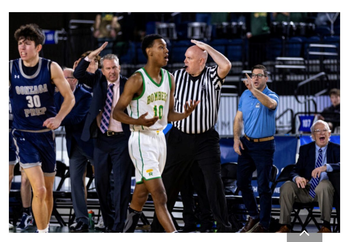 Basketball official whistling and signaling a foul as Gonzaga Prep and Bombers players and coaches react